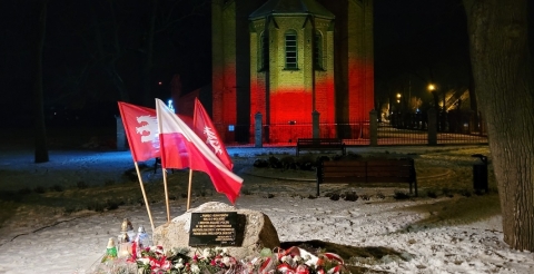 Na zdjęciu tablica upamiętniająca wybuch Powstania Wielkopolskiego z flagami powstańczymi i narodową. W tle biało-czerwona iluminacja na budynku dawnego kościoła ewangelickiego.
