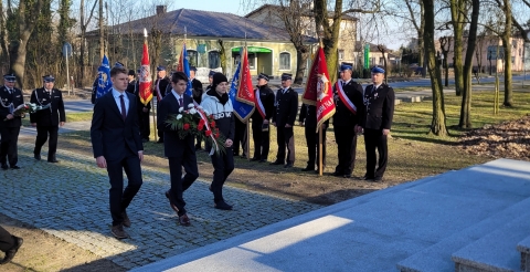 Delegacja Młodzieżowej Rady Gminy z wiązanką biało-czerwonych kwiatów zmierza w kierunku pomnika. W tle poczty sztandarowe Ochotniczych Straży Pożarnych ze sztandarami.