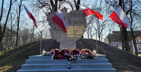 Pomnik Walk i Męczeństwa w Dusznikach udekorowany czterema biało-czerwonymi flagami na stojakach. U stóp pomnika leżą wiązanki biało-czerwonych kwiatów i znicze.