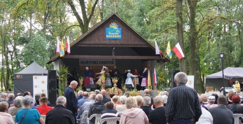 Obrzęd dożynkowy wykonywany przez zespół Poligrodzianie na scenie w parku w Dusznikach podczas Dożynek Gminnych 2021. Na pierwszym planie uczestnicy imprezy oglądający występ.